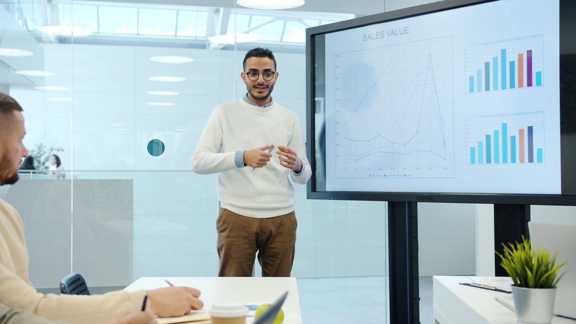 Analyst presenting sales charts on a large screen in a glass-walled meeting room
