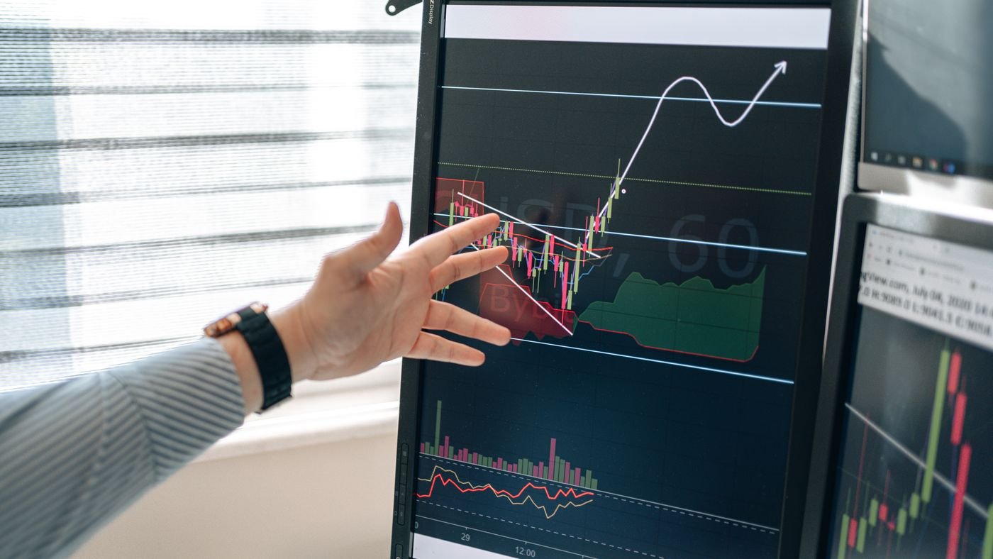 Hand pointing at a trend chart on a monitor beside a second data screen