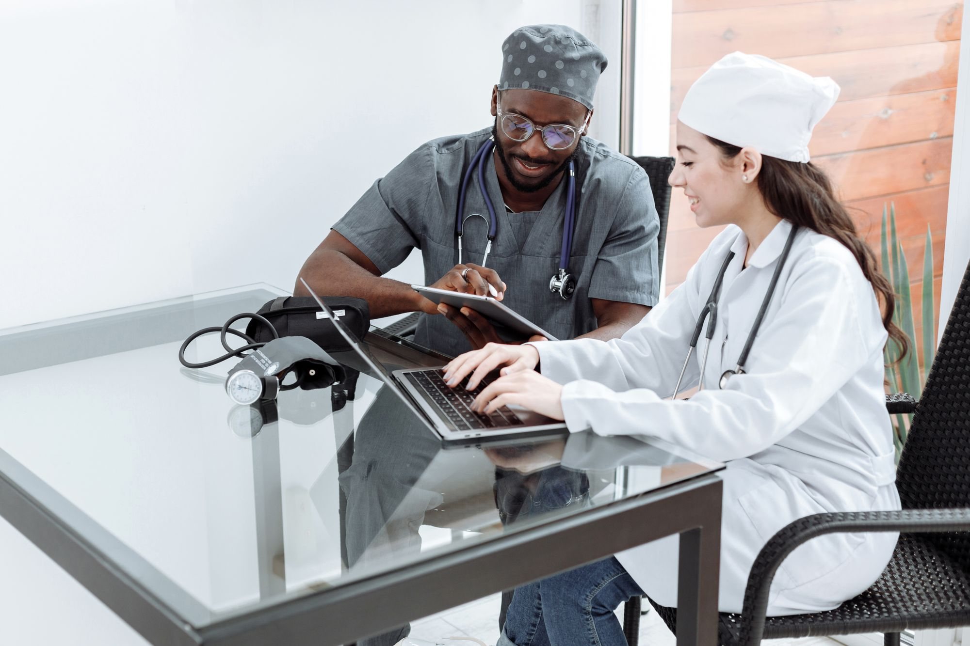Two doctors at a desk reviewing records on a laptop and a tablet